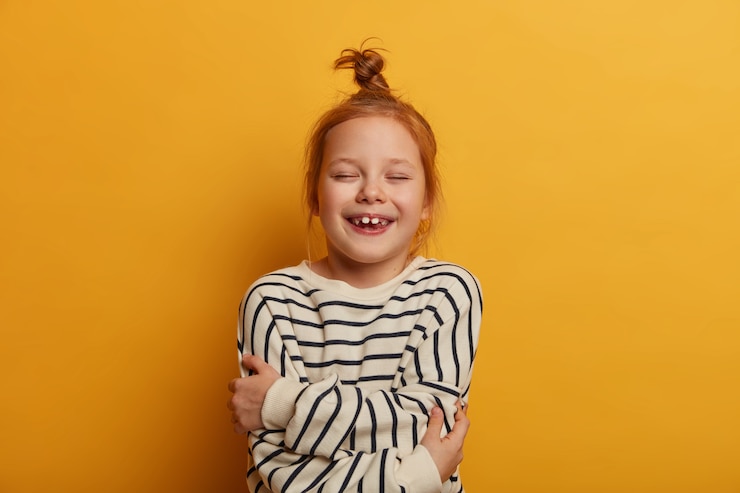 Photo gratuit l'enfant au gingembre s'embrasse, exprime son amour-propre, ferme les yeux du plaisir, porte un pull rayé, pose contre un mur jaune, se sent bien, montre des dents blanches, plein d'excitation