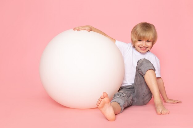 Un enfant blond vue de face en t-shirt blanc jouant avec une boule blanche ronde sur l'espace rose