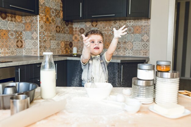 Enfant drôle debout dans une cuisine roustique jouant avec de la farine et le jetant en l'air.