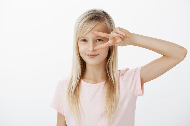 L'enfant énergique créatif veut danser le disco. Portrait de jeune fille blonde adorable intriguée en t-shirt rose, montrant la victoire ou le signe de la paix sur les yeux et souriant largement, ayant quelque chose à l'esprit