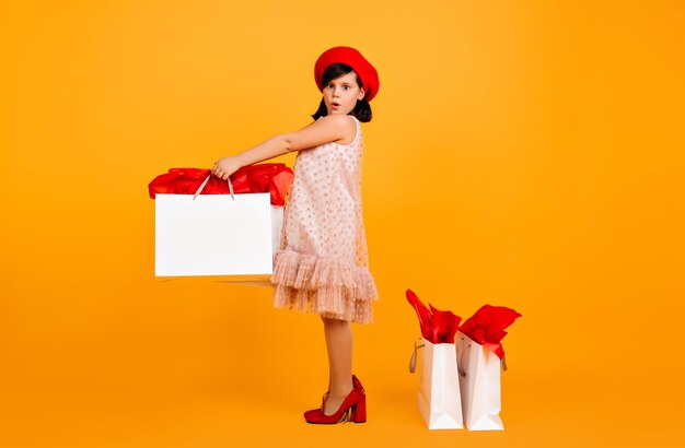 Enfant étonné en béret rouge et chaussures de maman debout sur fond jaune Prise de vue en studio d'un enfant mignon avec des sacs à provisions