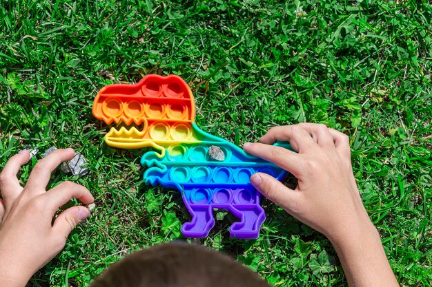 Enfant garçon jouer avec pop il sur l'herbe dans le parc. nouveau jouet de développement sensoriel en forme de dinosaure avec des bulles de silicone arc-en-ciel colorées. jeu de remuement à la mode, anti-stress et soulagement de l'anxiété.