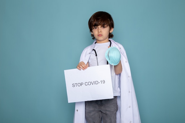 Photo gratuite enfant garçon tenant stop covid hashtag en combinaison médicale blanche et jeans gris sur mur bleu