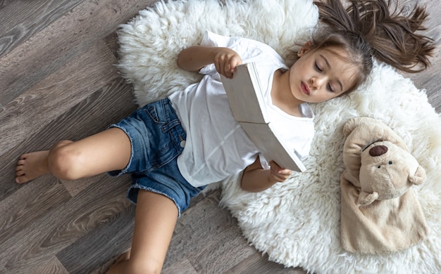 Photo gratuite l'enfant lit un livre allongé sur un tapis confortable à la maison avec son ours en peluche préféré.