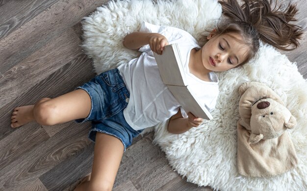 L'enfant lit un livre allongé sur un tapis confortable à la maison avec son ours en peluche préféré.