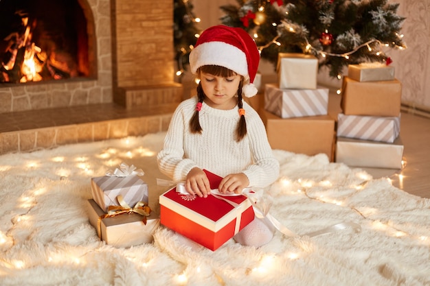 Enfant de sexe féminin optimiste portant un pull blanc et un chapeau de père Noël, ouvrant une boîte-cadeau, ayant une expression faciale concentrée, assis sur le sol près de l'arbre de Noël, des boîtes à cadeaux et une cheminée.