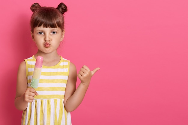 Enfant de sexe féminin tenant la crème glacée et pointant de côté avec le pouce, posant isolé sur un mur rose, gardant les lèvres arrondies, la petite fille a l'air pratique et drôle.