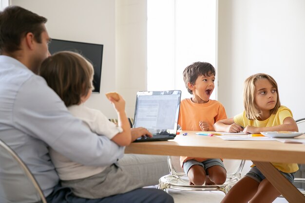 Des enfants heureux dessinent des griffonnages quand papa travaille sur un ordinateur portable et tient son fils sur les genoux. Enfants sortis de la peinture sur papier. Famille caucasienne assise à table. Concept d'enfance, de créativité et de week-end