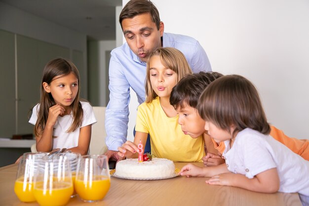 Enfants heureux et papa soufflant une bougie sur un gâteau et faisant un souhait. Jolie fille blonde célébrant son anniversaire avec des amis. Enfants heureux debout près de la table. Concept d'enfance, de célébration et de vacances