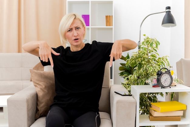 Ennuyé belle femme russe blonde est assise sur un fauteuil pointant vers le bas avec deux mains à l'intérieur du salon