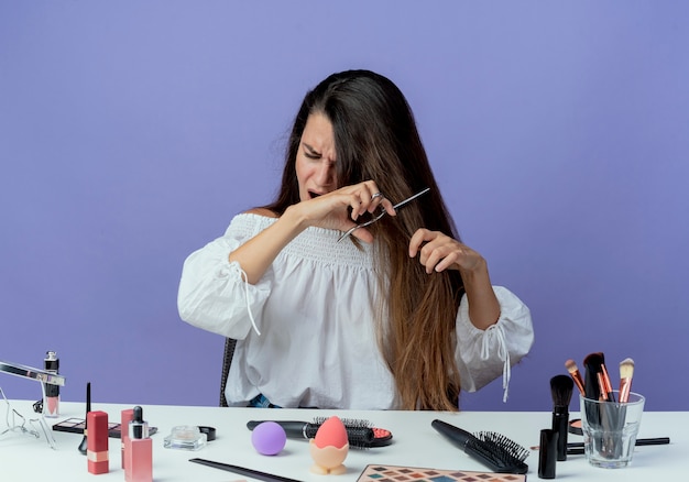 Ennuyé belle fille est assise à table avec des outils de maquillage fait semblant de couper les cheveux avec des ciseaux isolés sur un mur violet