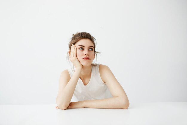 Ennuyé fatigué jeune jolie femme avec chignon pensant rêver assis à table sur fond blanc.