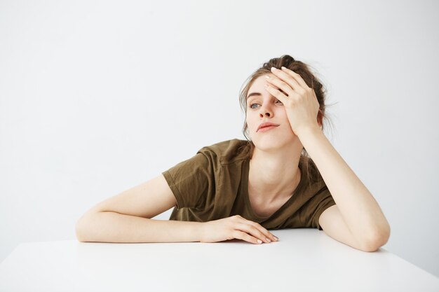 Ennuyé fatigué terne jeune femme étudiante avec chignon assis à table sur fond blanc.