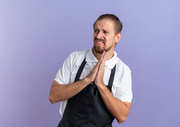 Ennuyé jeune beau coiffeur portant l'uniforme mettant les mains dans le geste de prier à côté isolé sur violet avec copie espace