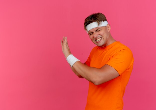 Ennuyé jeune bel homme sportif portant bandeau et bracelets debout en vue de profil gesticulant non avec les yeux fermés isolé sur rose avec espace copie