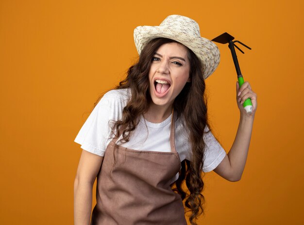 Ennuyé jeune femme jardinière en uniforme portant chapeau de jardinage détient houe râteau isolé sur mur orange avec copie espace