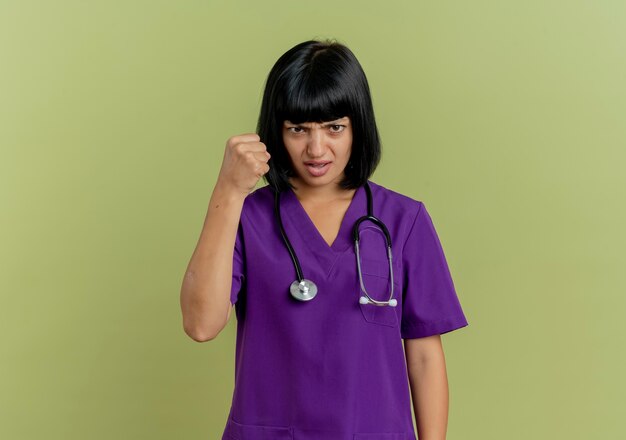 Ennuyé jeune femme médecin brune en uniforme avec stéthoscope garde le poing regardant la caméra isolée sur fond vert olive avec copie espace