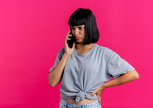 Ennuyé jeune fille brune caucasienne met la main sur la taille en parlant au téléphone à côté isolé sur fond rose avec espace de copie