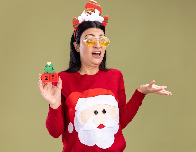 Ennuyé jeune fille caucasienne portant bandeau de père Noël et pull avec des lunettes tenant jouet arbre de Noël avec date regardant côté montrant la main vide isolé sur mur vert olive