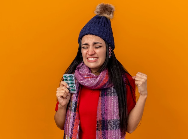 Ennuyé jeune fille malade de race blanche portant un chapeau d'hiver et une écharpe tenant un pack de capsules avec d'autres packs sous chapeau serrant le poing avec les yeux fermés isolé sur un mur orange avec espace de copie