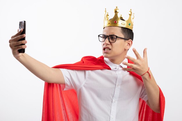 Ennuyé jeune garçon de super-héros en cape rouge portant des lunettes et une couronne en gardant la main dans l'air en prenant selfie isolé sur fond blanc