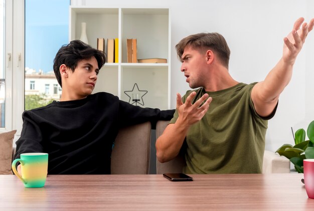 Ennuyé jeune homme beau blond pointe sur le côté avec deux mains assis à table et regardant jeune beau garçon brune à l'intérieur de la salle de séjour