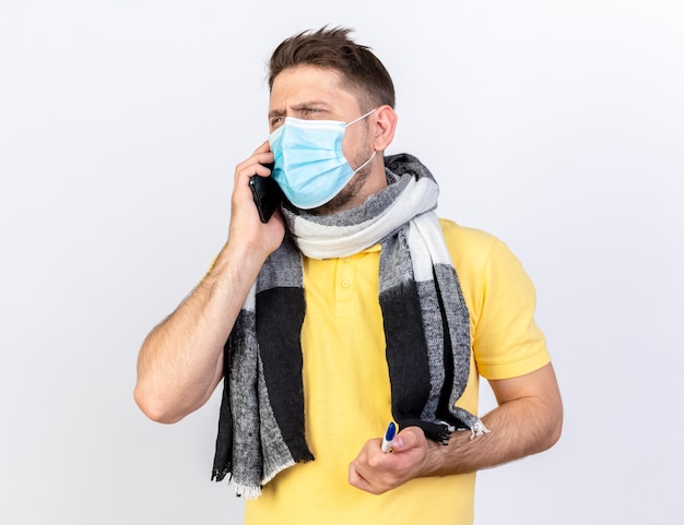 Ennuyé jeune homme malade blonde portant un masque médical et un foulard parle au téléphone détient un thermomètre isolé sur un mur blanc