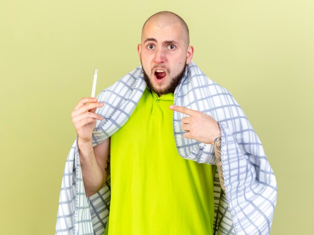 Photo gratuite ennuyé jeune homme malade de race blanche enveloppé dans des prises à carreaux et des points au thermomètre isolé sur un mur vert olive avec espace de copie