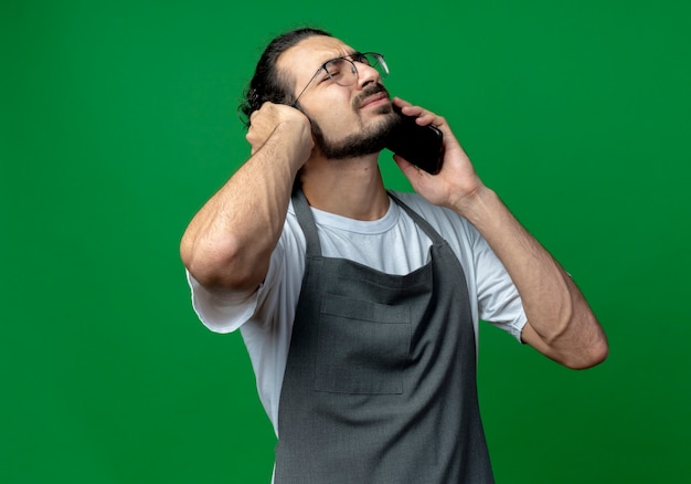 Ennuyé jeune homme de race blanche coiffeur portant l'uniforme et des lunettes de parler au téléphone avec la main sur l'oreille et les yeux fermés isolés sur fond vert avec espace copie