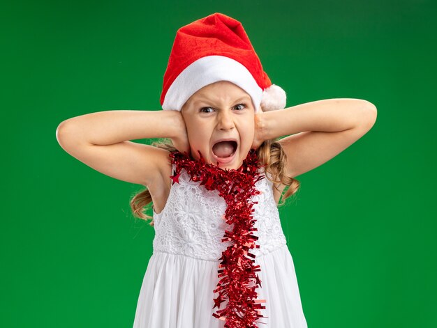 Ennuyé petite fille portant un chapeau de Noël avec guirlande sur les oreilles couvertes de cou isolé sur mur vert