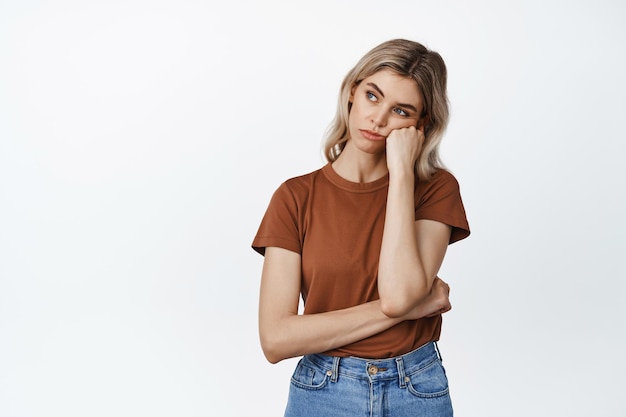 Ennuyé et triste fille regardant la tête penchée à gauche sur le poing avec une expression de visage sceptique de mauvaise humeur debout sur fond blanc