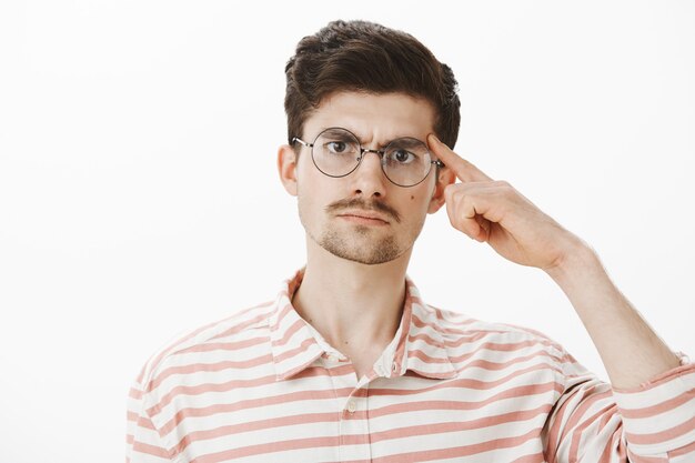 Enseignant ennuyé ennuyé demandant de penser avec le cerveau. Homme sérieux mécontent avec barbe et moustache dans des lunettes à la mode, tenant l'index sur la tempe et gronder étudiant sur mur gris