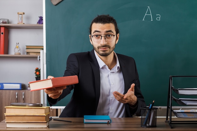 Enseignant heureux portant des lunettes tenant un livre, assis à table avec des outils scolaires en classe
