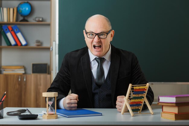 Un enseignant portant des lunettes avec un registre de classe assis au bureau de l'école devant le tableau noir dans la salle de classe à l'aide d'abaque en colère et frustré en serrant les poings en criant