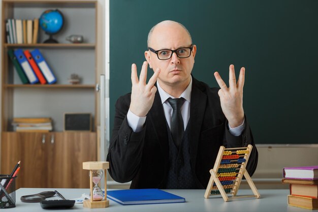 Enseignant portant des lunettes avec registre de classe assis au bureau de l'école devant le tableau noir dans la salle de classe à l'aide d'abaque montrant le numéro six avec les doigts avec une expression sérieuse