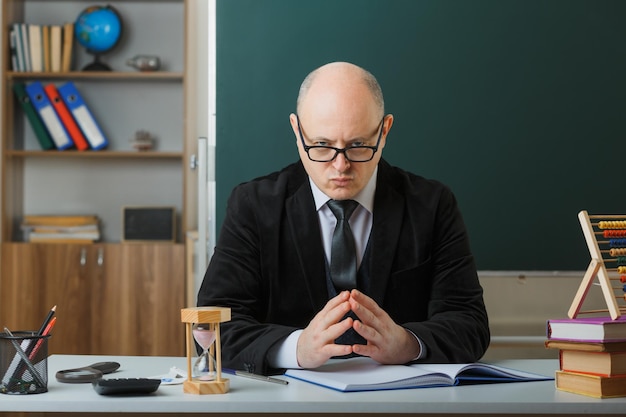 Enseignant portant des lunettes vérifiant le registre de classe regardant la caméra avec un visage sérieux tenant les paumes ensemble attendant quelque chose assis au bureau de l'école devant le tableau noir dans la salle de classe