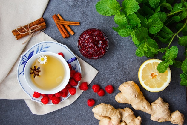 Photo gratuite ensemble de citron, framboises et confiture de framboises dans des soucoupes, gingembre, feuilles de menthe, cannelle sèche et une tasse de thé à la camomille sur un chiffon