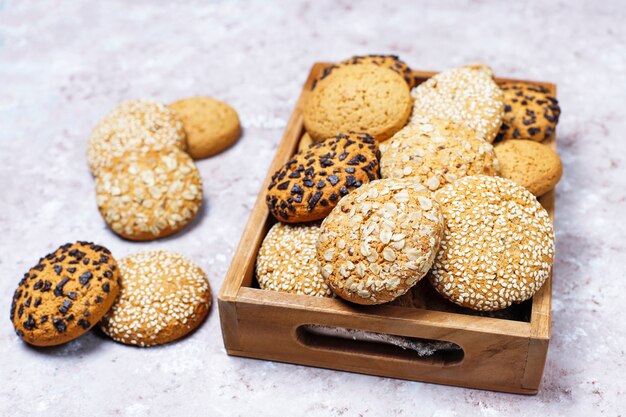 Ensemble de divers biscuits de style américain dans un plateau en bois sur fond de béton clair. Sablés aux graines de sésame, beurre d'arachide, flocons d'avoine et biscuits aux pépites de chocolat.