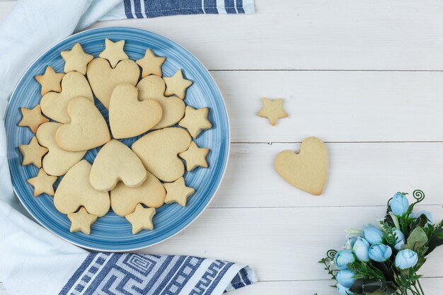 Ensemble de fleurs et de biscuits dans une assiette sur fond de serviette en bois et de cuisine. pose à plat.