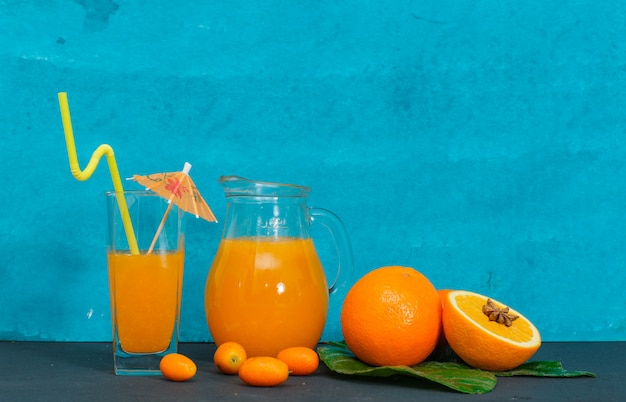 Photo gratuite ensemble de jus dans des verres, des feuilles et des oranges sur une surface texturée cyan. vue de côté.