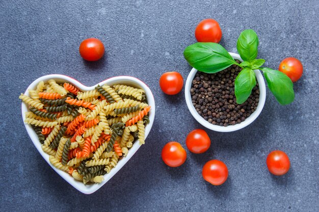 Ensemble de tomates, poivre noir, feuilles et pâtes macaroni colorées dans un bol en forme de coeur sur une surface grise. vue de dessus.