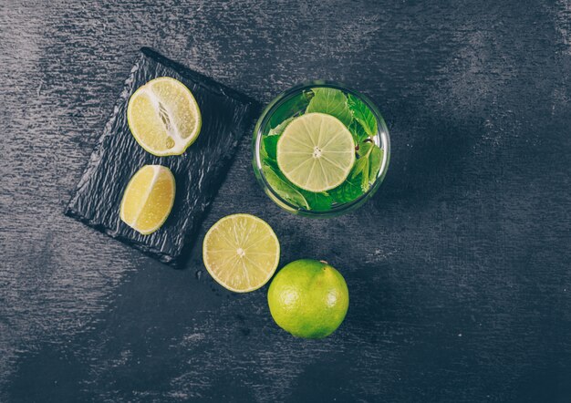 Ensemble de tranches et de citrons verts dans un verre d'eau sur un fond texturé noir. vue de dessus.