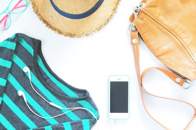 Ensemble de vêtements et d&#39;accessoires féminins à plat et accessoires avec un t-shirt, des lunettes de mode, un chapeau avec un téléphone portable et un écouteur sur fond blanc.