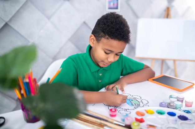 Enthousiasme. Garçon aux cheveux bouclés à la peau foncée en t-shirt vert esquissant avec enthousiasme une photo avec un crayon de couleur assis à table dans la salle lumineuse