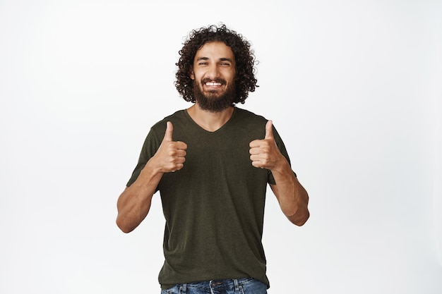 Enthousiaste bel homme souriant montrant les pouces vers le haut approuver et comme debout dans des vêtements décontractés sur fond blanc