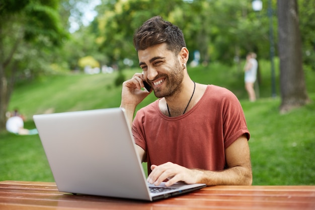 Enthousiaste bel homme utilisant un ordinateur portable dans le parc et parler au téléphone heureux
