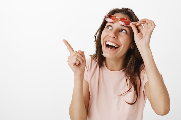 Enthousiaste belle fille avec des lunettes de soleil posant contre le mur blanc