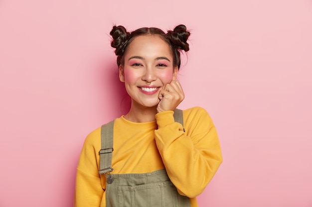 Enthousiaste belle jeune femme asiatique aux joues rouges, garde une main sous le menton, a deux petits pains, porte un pull jaune et une salopette marron