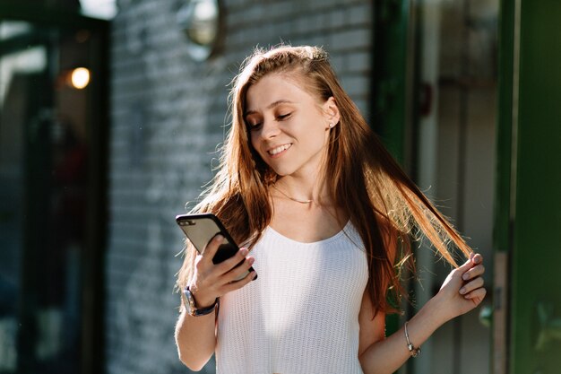 Enthousiaste belle jeune femme aux longs cheveux brun clair à l'aide de smartphone tout en marchant sur la rue ensoleillée et joue avec ses cheveux.