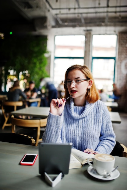 Enthousiaste belle jeune femme rousse dans des verres à l'aide de son pavé tactile de téléphone et de son ordinateur portable alors qu'elle était assise sur son lieu de travail au café avec une tasse de café